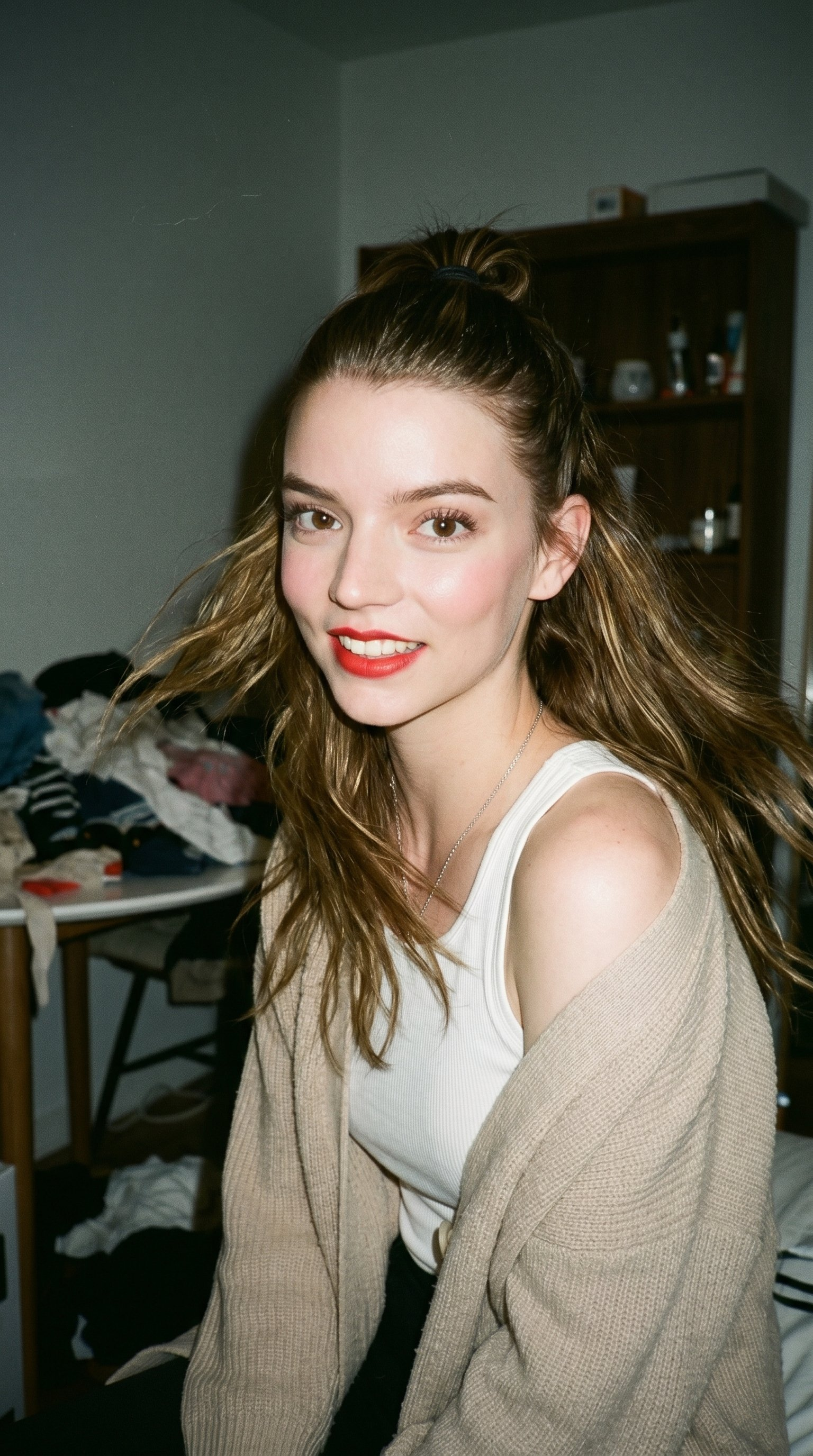 A woman with long, wavy brown hair in a messy bun, wearing a beige knit cardigan, smiling softly against a cluttered room backdrop with soft lighting.