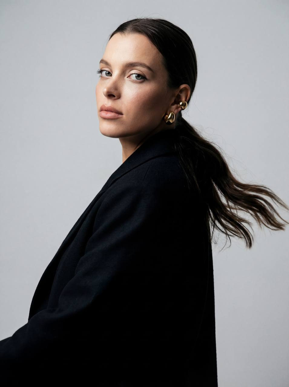 A photorealistic fashion portrait featuring a person in a sleek low ponytail, wearing a straight-cut blazer and large ear cuffs.
