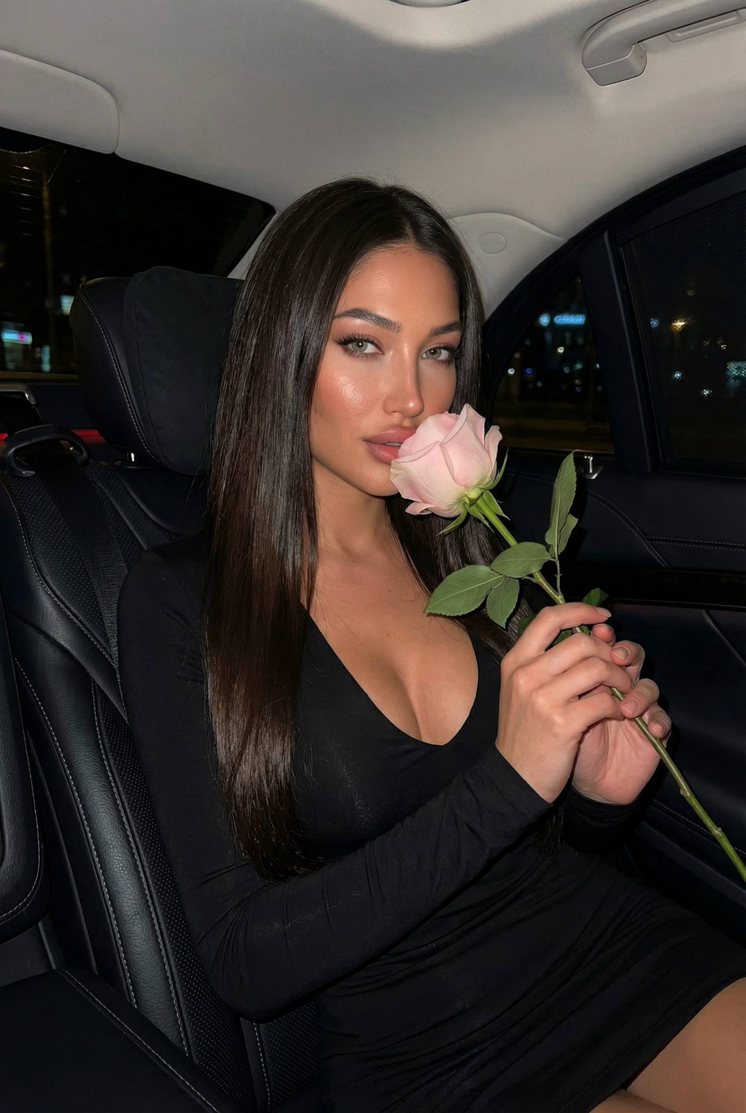 A woman in a fitted black dress seated in a car, holding a pale pink rose, gazing softly at the camera.