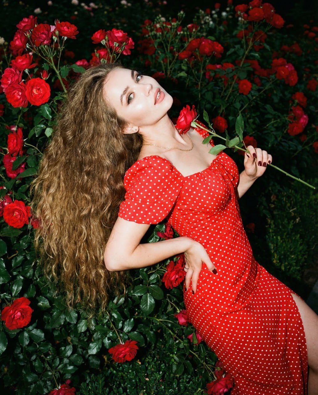 A woman in a fitted red dress surrounded by blooming red roses, lying gracefully with a playful gaze.