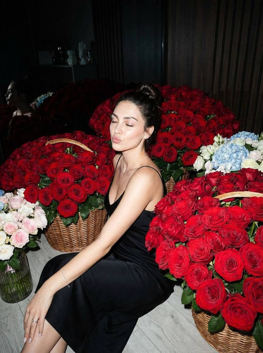 A woman in an elegant black dress surrounded by lush red roses, eyes closed, showcasing a serene atmosphere.