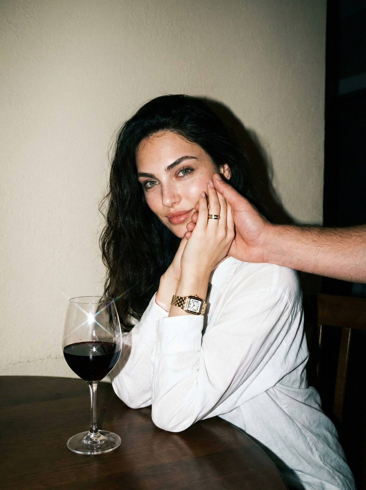 A woman sitting at a table in a cozy setting, illuminated with direct flash creating sharp shadows, softly smiling with her hand on her cheek.