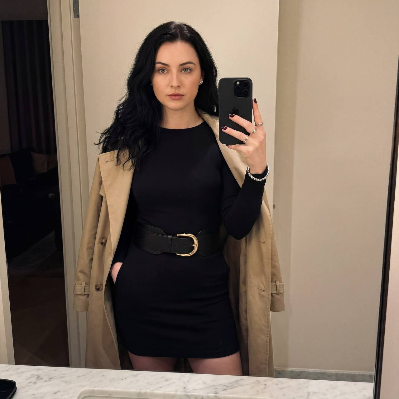 A stylish woman takes a mirror selfie holding an iPhone 16 Pro Max, wearing a mini-dress with a beige trench coat and accessories.