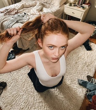A woman with vibrant reddish-orange hair tying her ponytail, kneeling on a white rug in a messy bedroom with natural light.