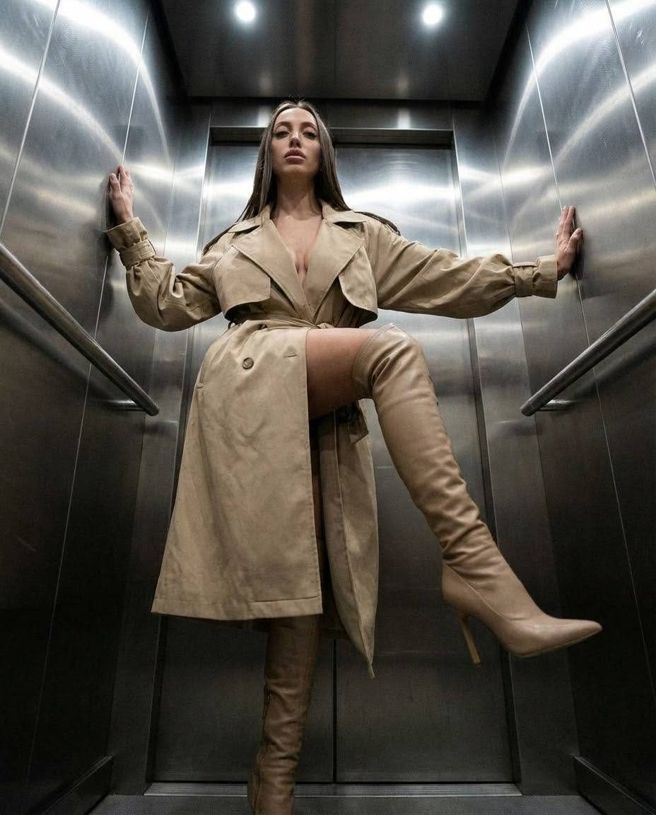 A woman in a trench coat poses dramatically in an elevator, leaning against chrome walls with a raised leg and hands pressed against the doors.