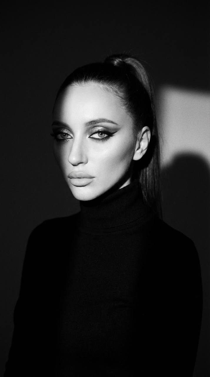 Dramatic waist-up portrait of a woman in a dimly lit studio, wearing a cashmere turtleneck with slicked-back hair and bold makeup.