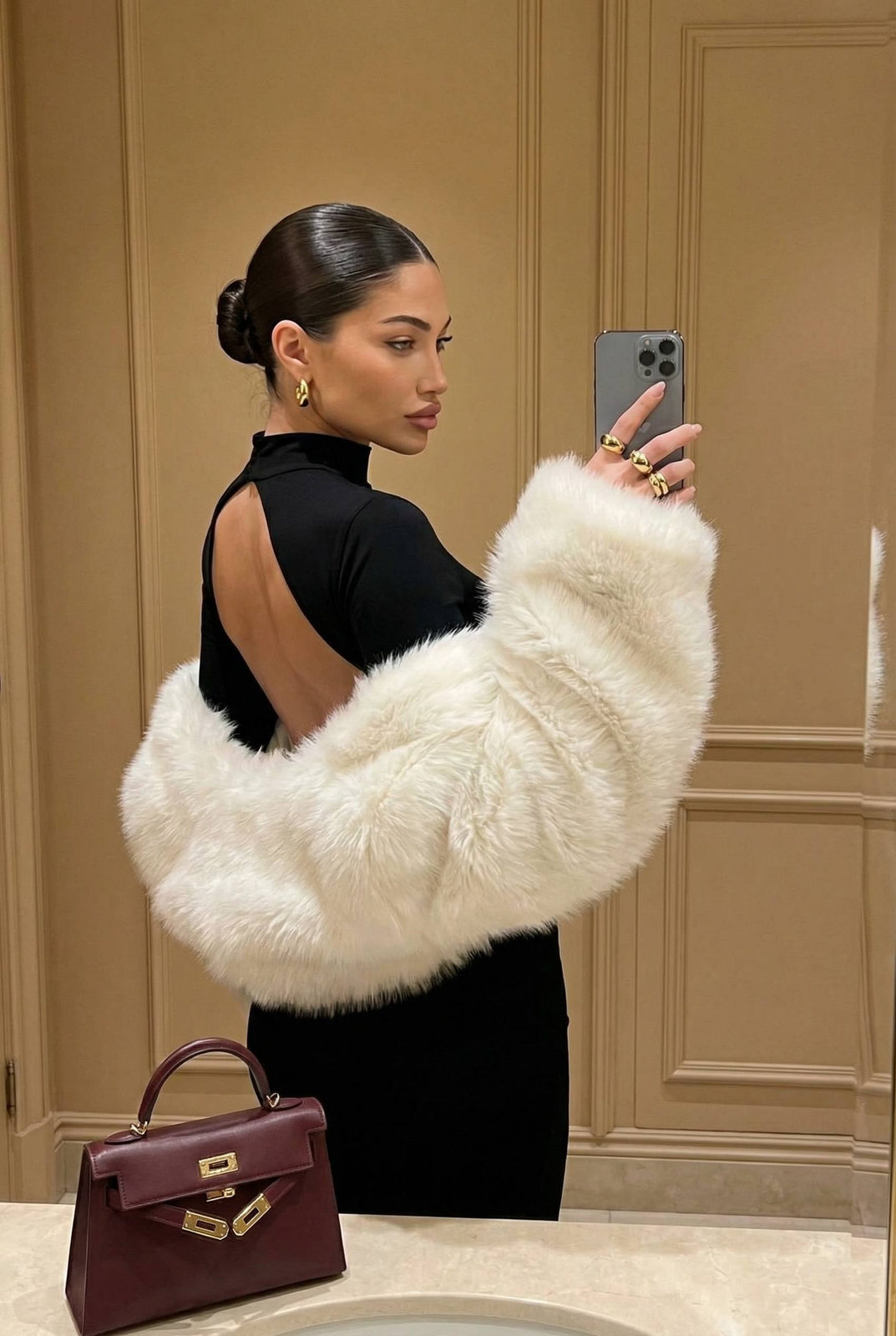 A woman taking a mirror selfie in a sleek black dress with a faux-fur stole, showcasing an elegant interior.