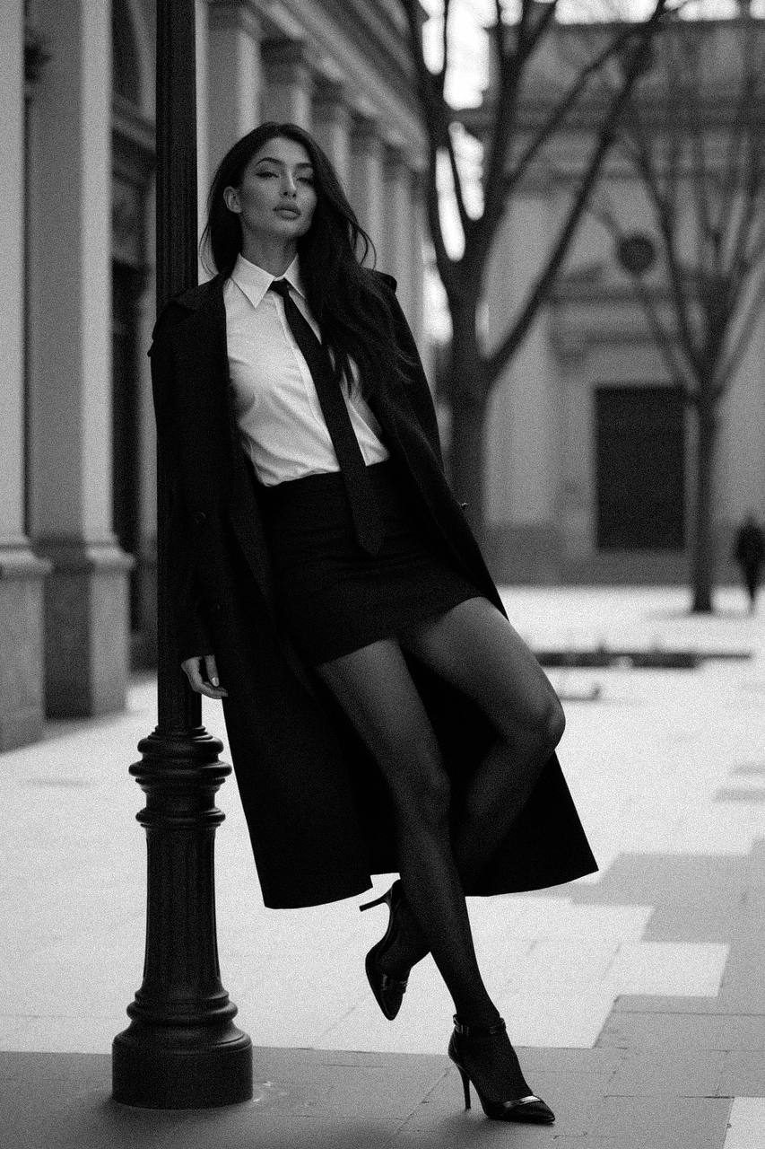 A high-fashion woman in a crisp button-up shirt and oversized coat leaning against a lamppost in an urban setting.