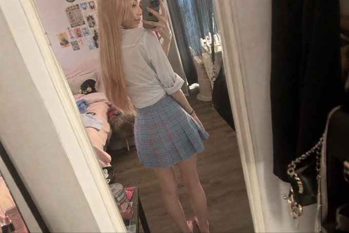 A woman taking a mirror selfie in a white shirt and plaid skirt, reflecting in full length with soft indoor lighting.