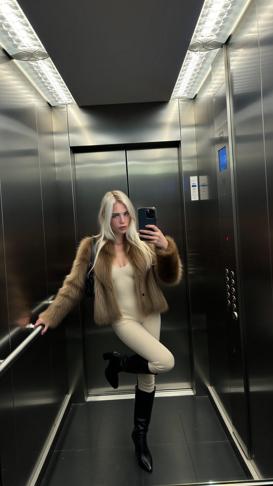 A confident woman taking a selfie in a modern elevator, showcasing her brown plush fur jacket and cream jumpsuit.