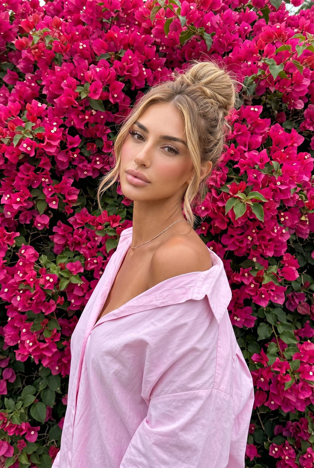 Woman in pastel pink shirt standing in front of vivid magenta-pink bougainvillea flowers, looking softly at camera.