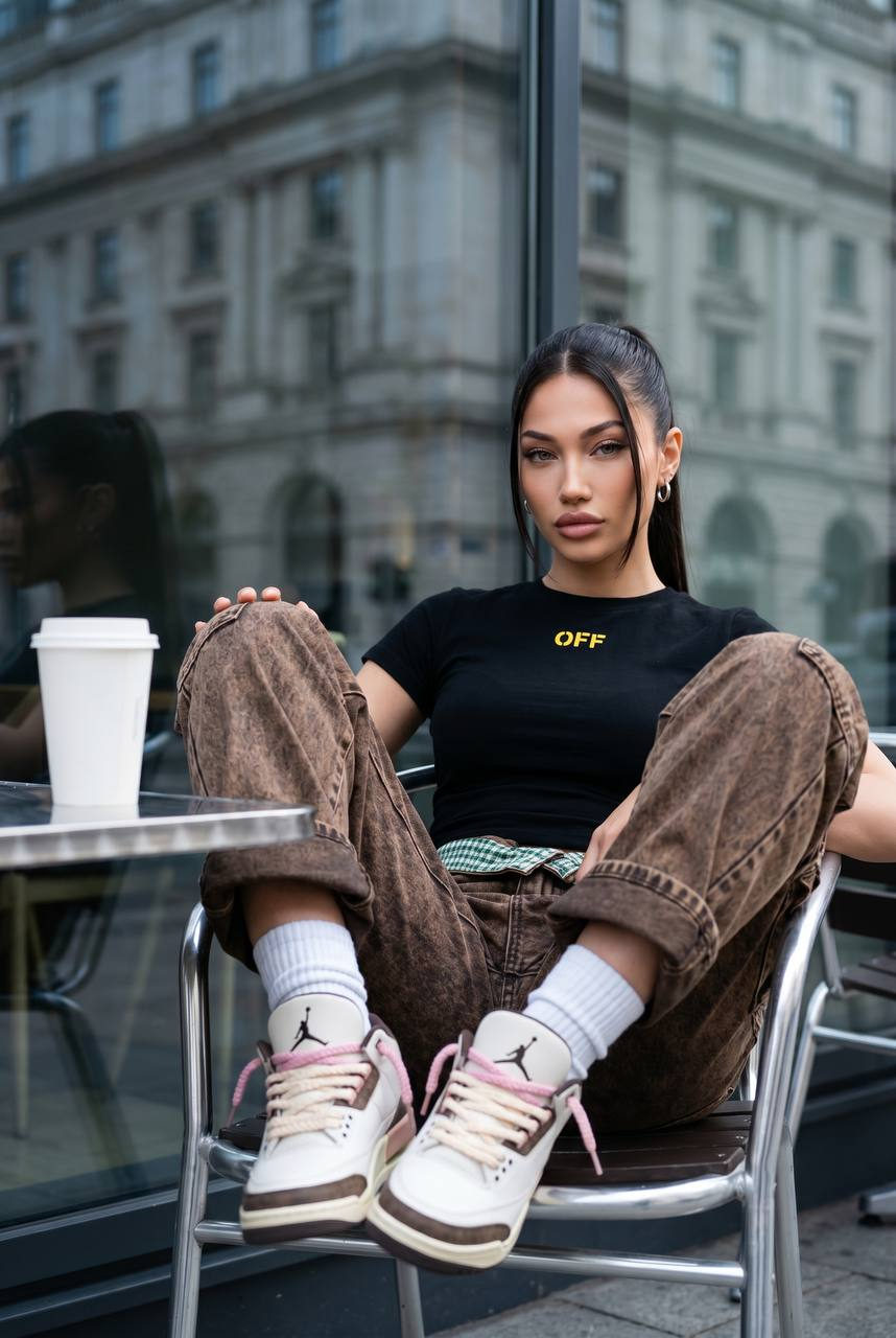 Photorealistic full-body portrait of a woman sitting relaxed at a cafe table with a white coffee cup, leaning back, sneaker-clad feet visible.