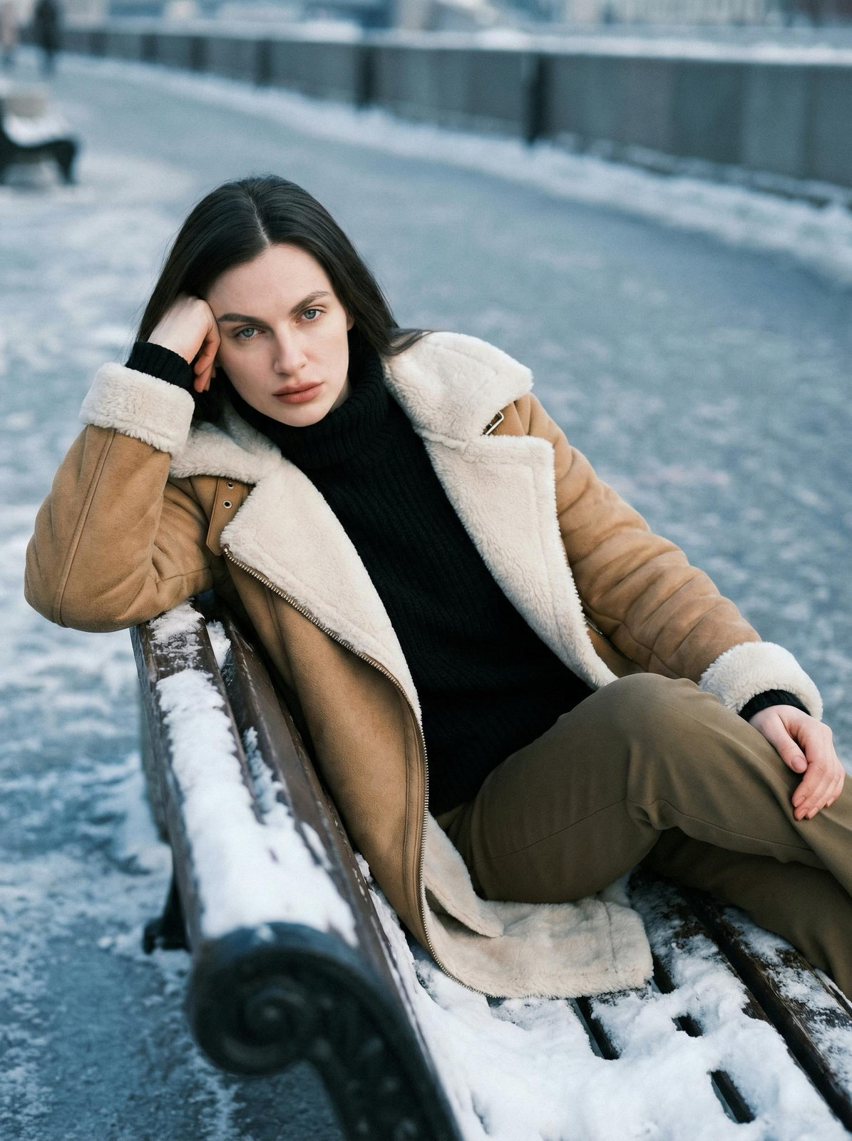 A woman in a caramel-beige coat reclining on a snowy bench in winter, with brown makeup and frosty surroundings.