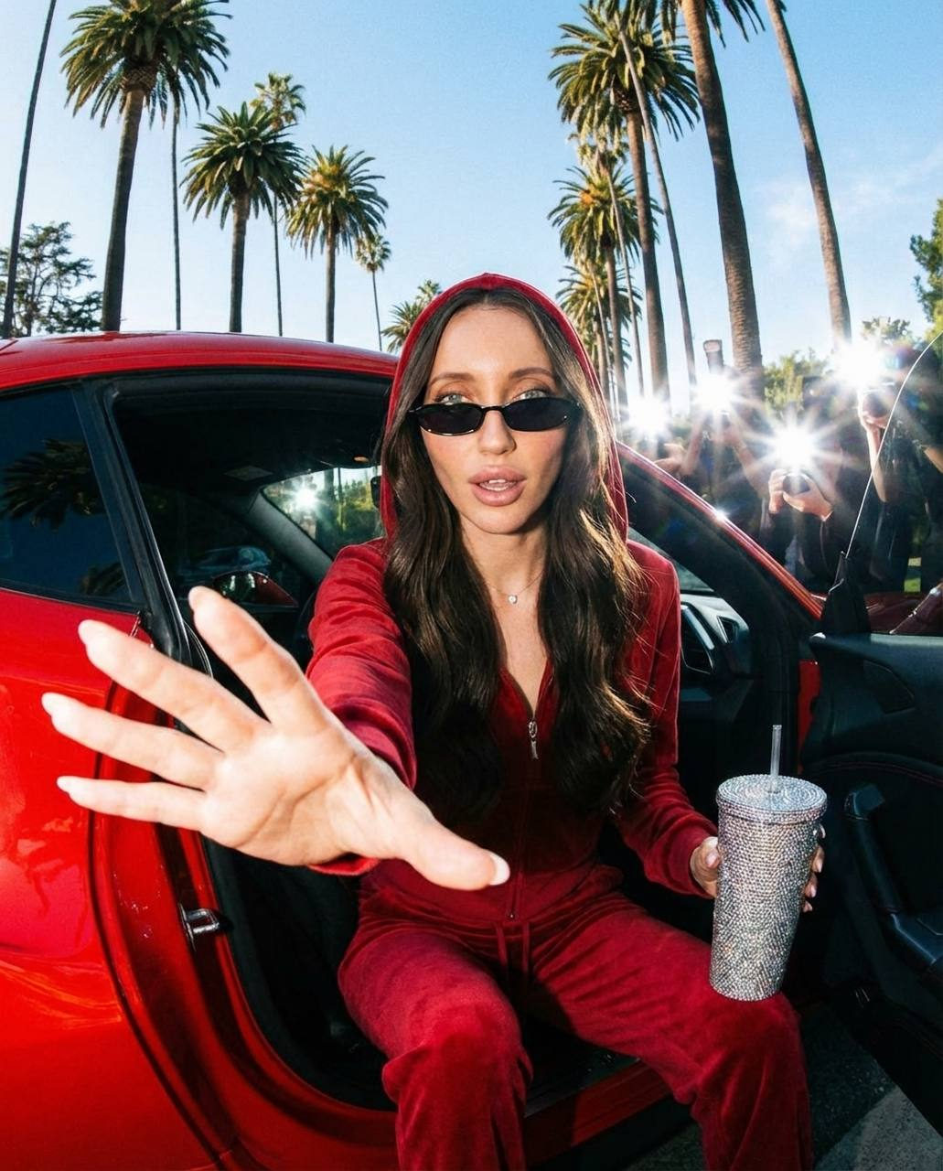 A glamorous woman in sunglasses sitting in a red car door, palm trees in the background.