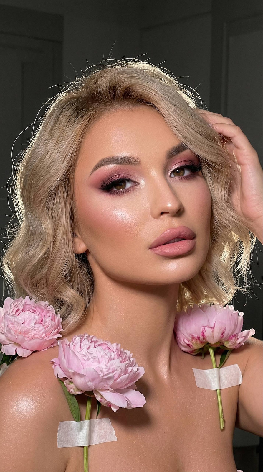 A close-up portrait of a woman with pink smoky eyeshadow and glowing skin, framed by delicate peonies.