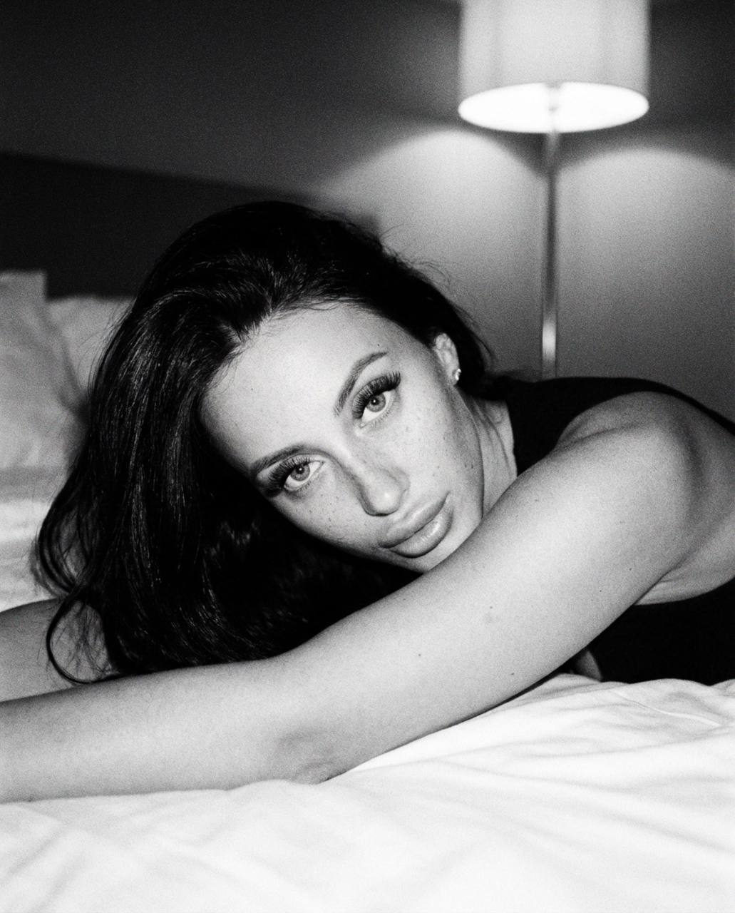 Close-up shot of a girl on a white bed, black and white with grain, confidently gazing at the camera.
