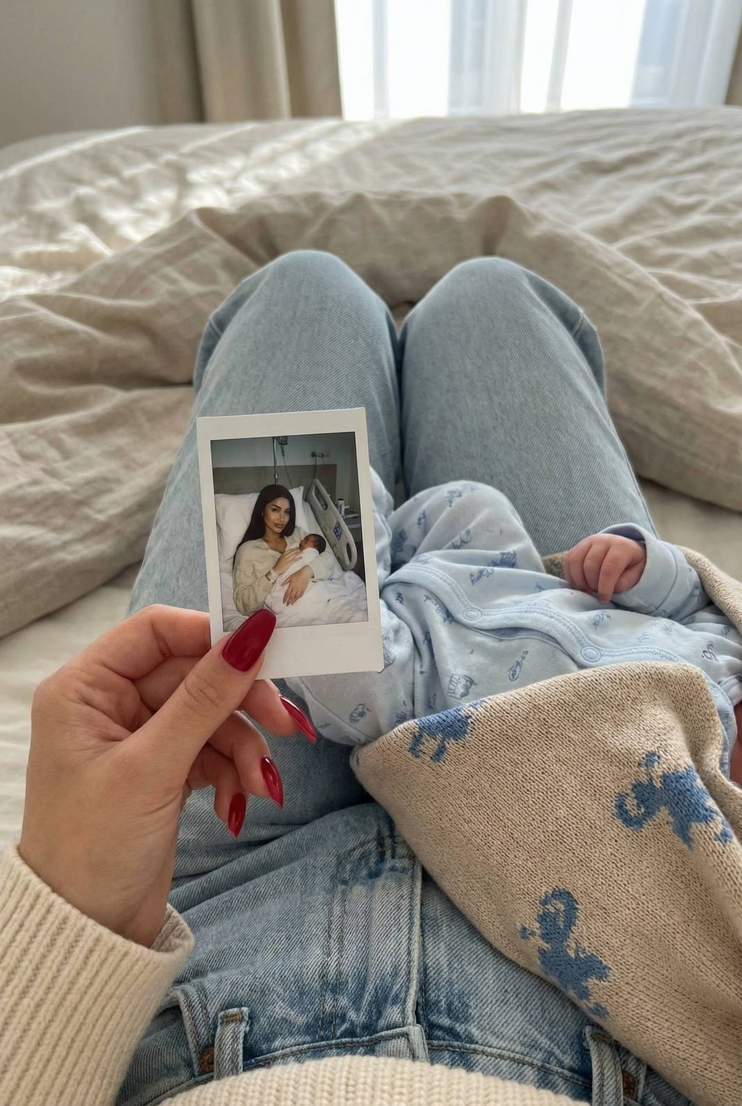 Close-up shot of a woman holding a polaroid photo with a baby resting against her legs on a bed.