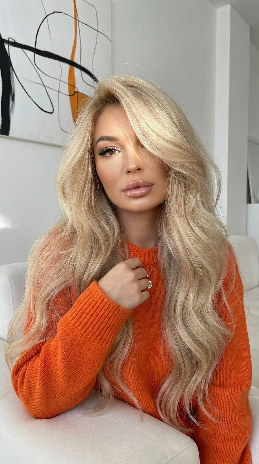 A woman in a cozy bright orange sweater sitting on a sofa, adjusting a pendant, with soft light illuminating her face.