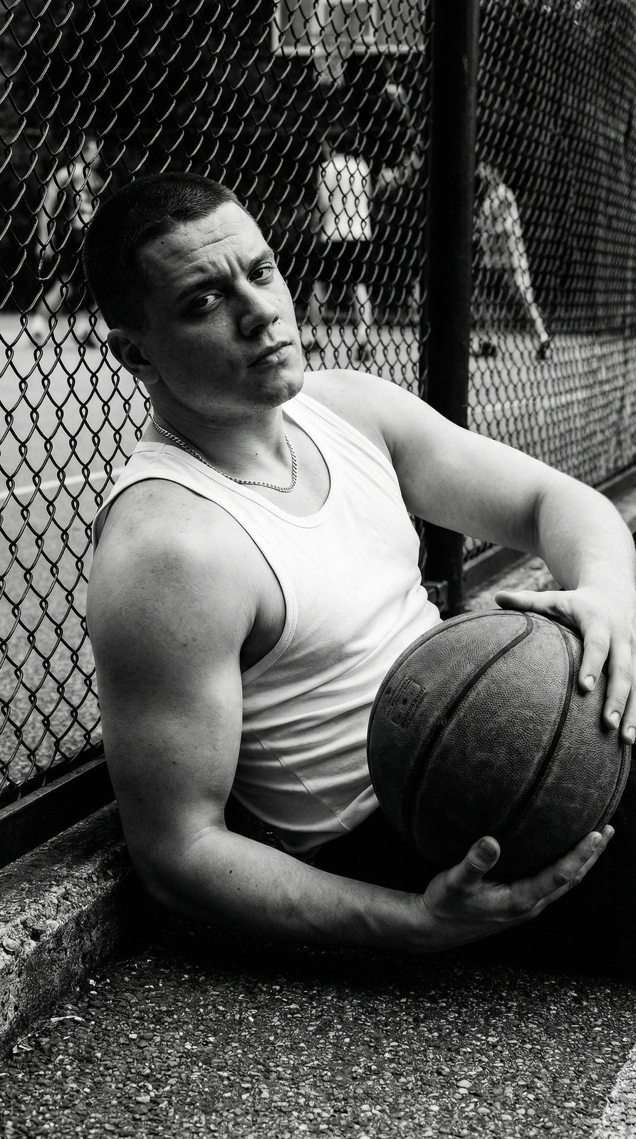 A muscular person sitting against a basketball court fence, relaxed with a basketball, gazing intently at the camera.