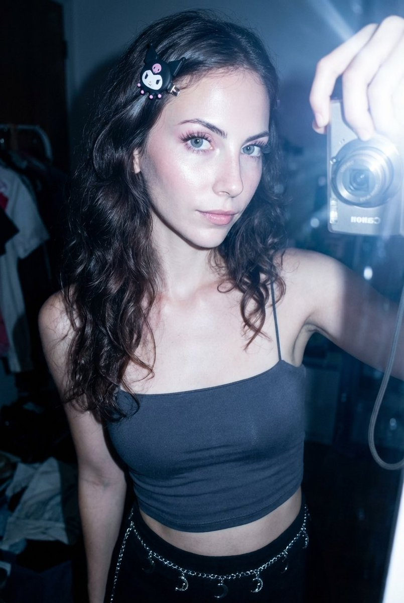 A teenage girl with dark brown wavy hair and a Sanrio Kuromi clip, wearing a dark grey crop top, showcasing a glowing radiant complexion.