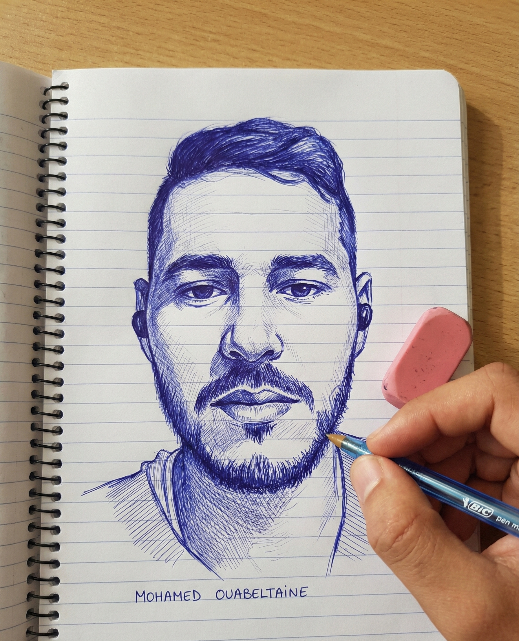 A detailed ink drawing of a person with intricate facial features, holding a pen over a notebook page.