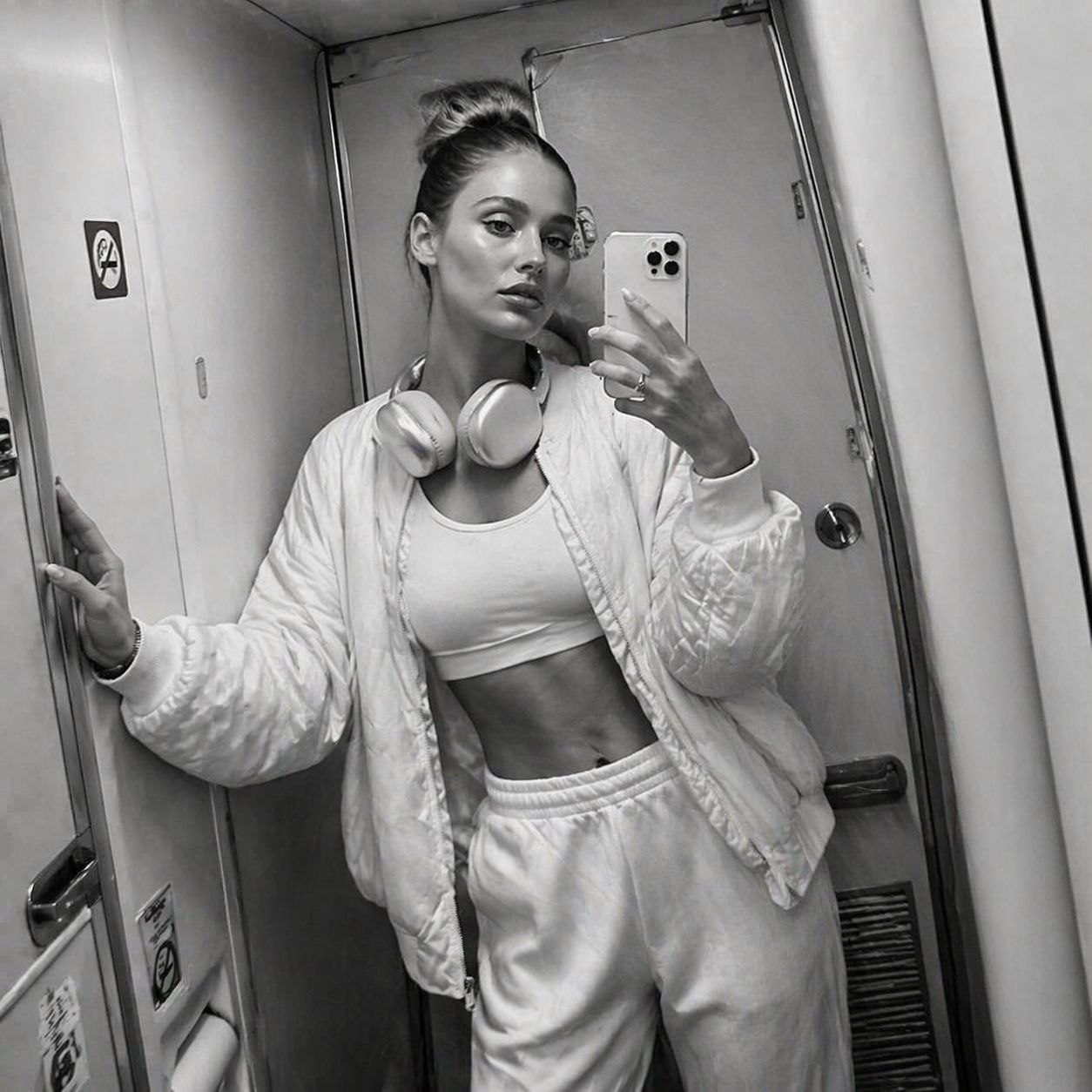 A person takes a mirror selfie in an airplane restroom, showcasing a unique asymmetrical pose and a white oversized outfit.