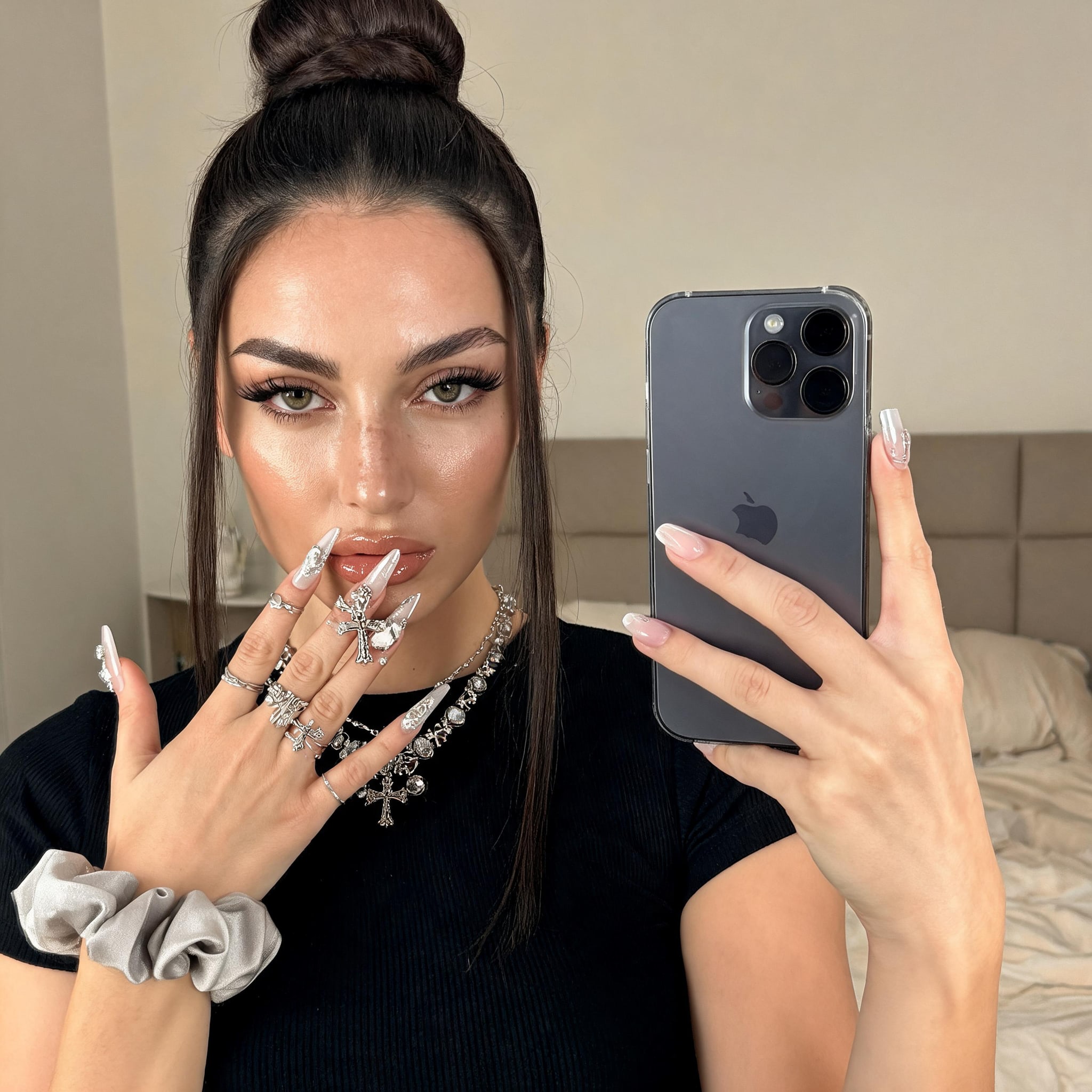 A woman taking a selfie in a mirror, showcasing nude makeup with radiant skin, a black t-shirt, and silver jewelry in a cozy bedroom.