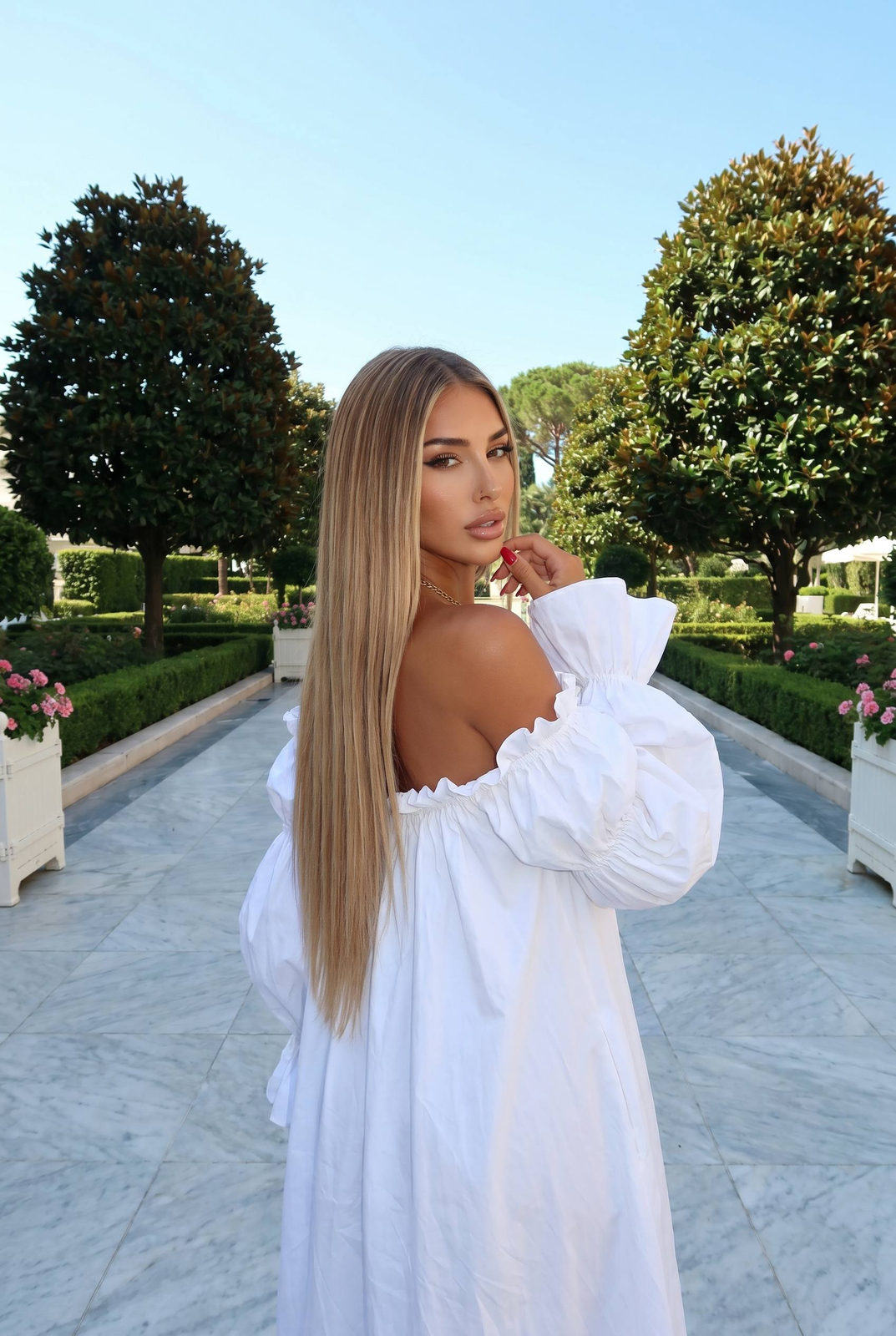 A woman stands on a wide marble-tiled promenade, wearing an oversized white dress, with long straight hair and surrounded by lush greenery.