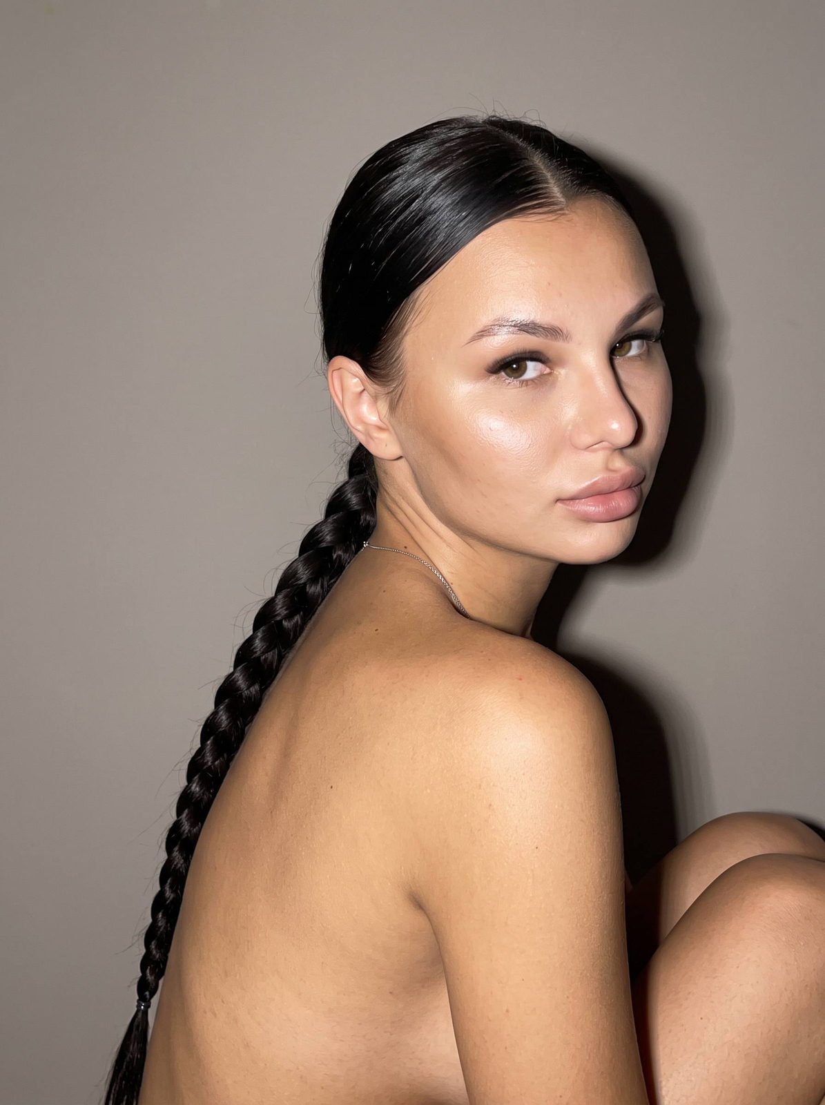 Close-up portrait of a woman with sleek hair and bare shoulders, sitting indoors against a warm-grey wall, illuminated by strong flash.