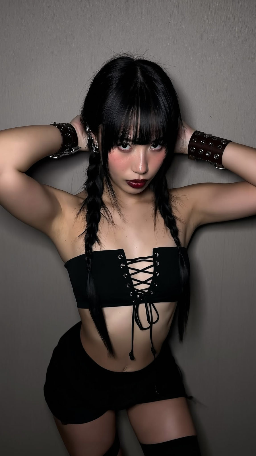 Close-up portrait of a woman with braids and intense gaze, wearing a bandeau top against a gray wall.