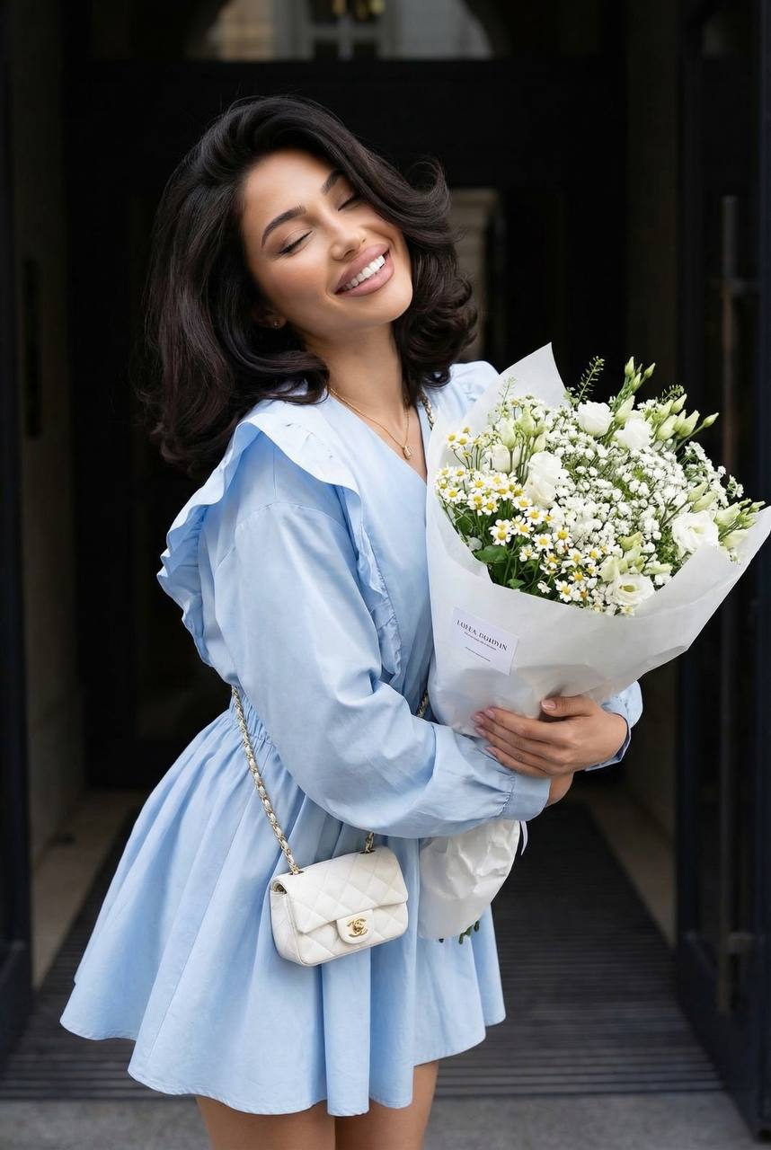 Woman in a pastel blue dress holding a bouquet of white flowers outdoors with dark architecture in the background.