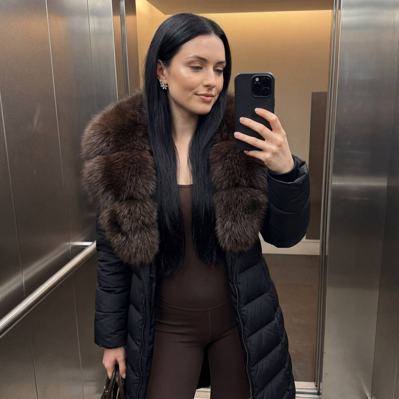 Ultra-realistic selfie in an elevator featuring a woman in a long quilted jacket with a fluffy dark brown fur collar and an iPhone in hand.