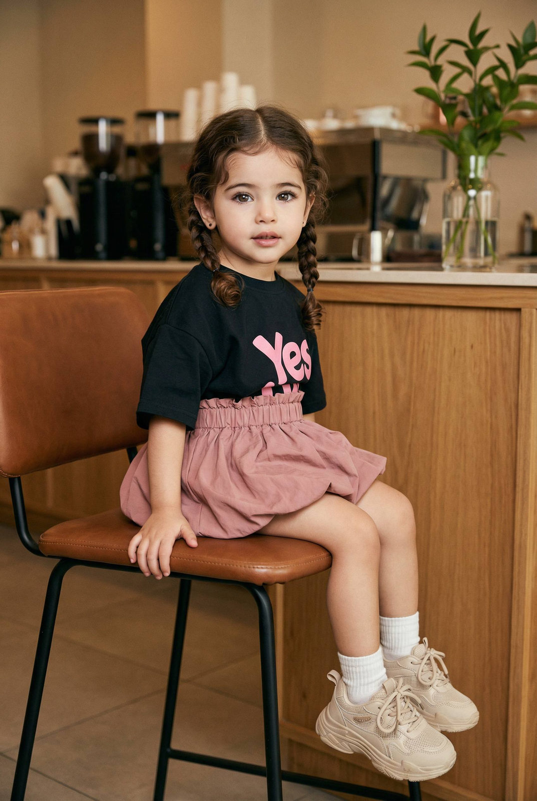 A little girl sitting sideways on a leather café chair, relaxed pose, wearing a pink t-shirt and skirt with blurred café background.