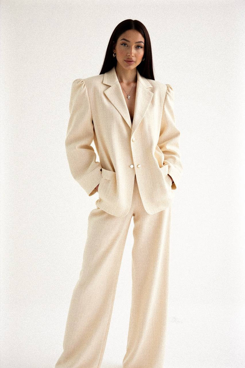 Woman in oversized cream checkered suit standing confidently with hands in pockets, showcasing high-fashion style in bright daylight with soft shadows.