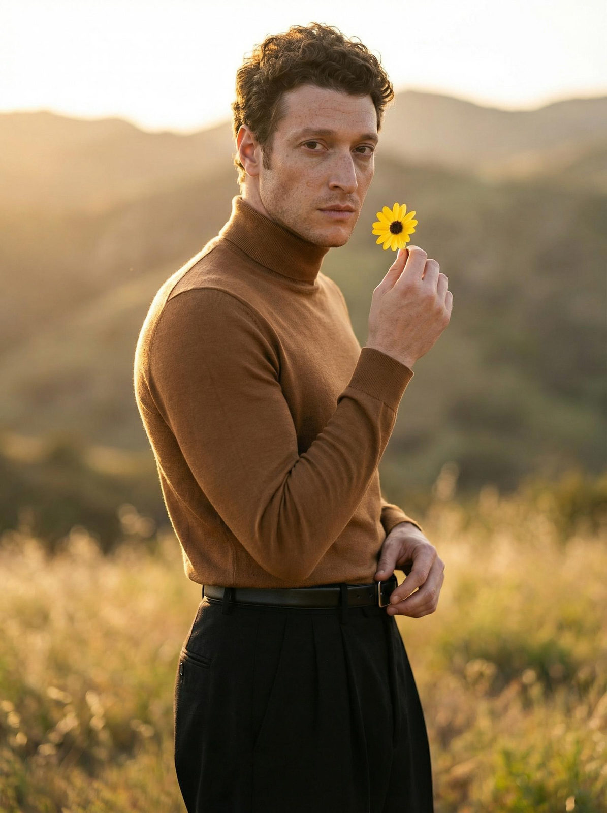 A man poses in a sunset landscape, holding a small yellow flower with backlighting enhancing his silhouette.