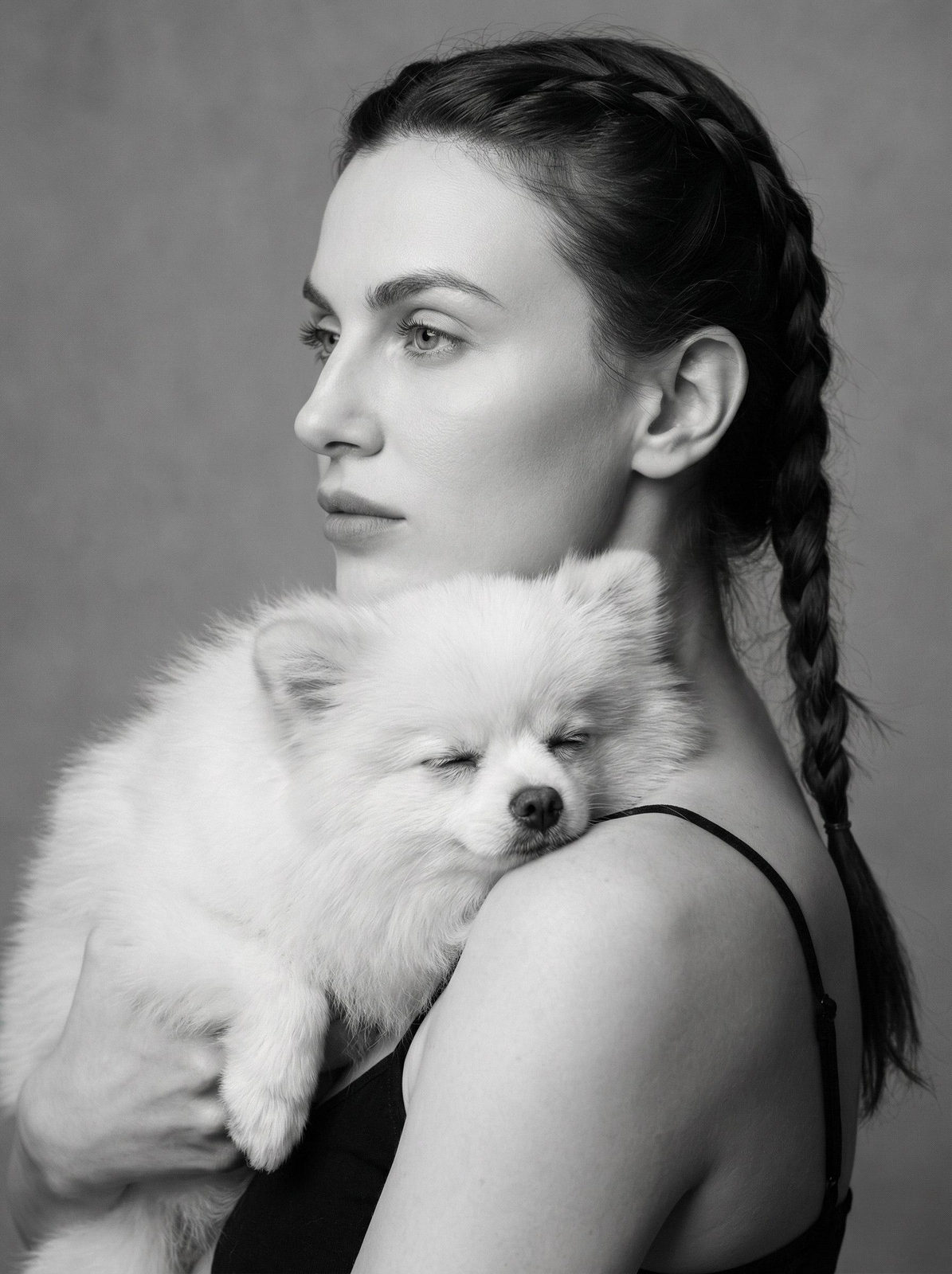 A woman in profile with braided pigtails holds a dog, showcasing soft lighting and a minimalist gray background.