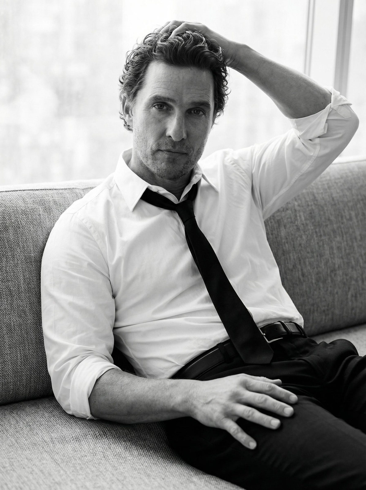 A relaxed man seated on a textured sofa, hand in hair, soft lighting casting gentle shadows on his face.