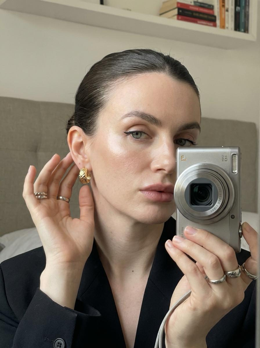 A woman taking a selfie in front of a mirror, holding a compact camera, showcasing her earrings and stylish black jacket.