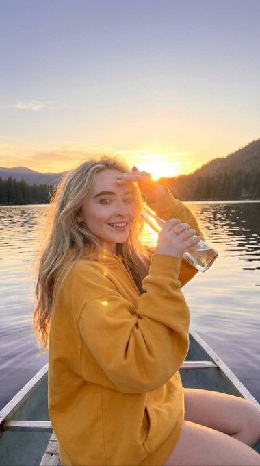 A woman in a mustard yellow hoodie sits in a canoe on a calm lake at sunset, holding a glass bottle and shielding her eyes from the sun.