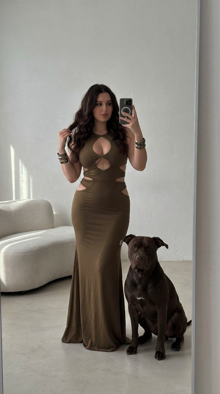 Full-body shot of a woman wearing a form-fitting olive-brown evening dress, taking a mirror selfie next to a large dark-brown dog.