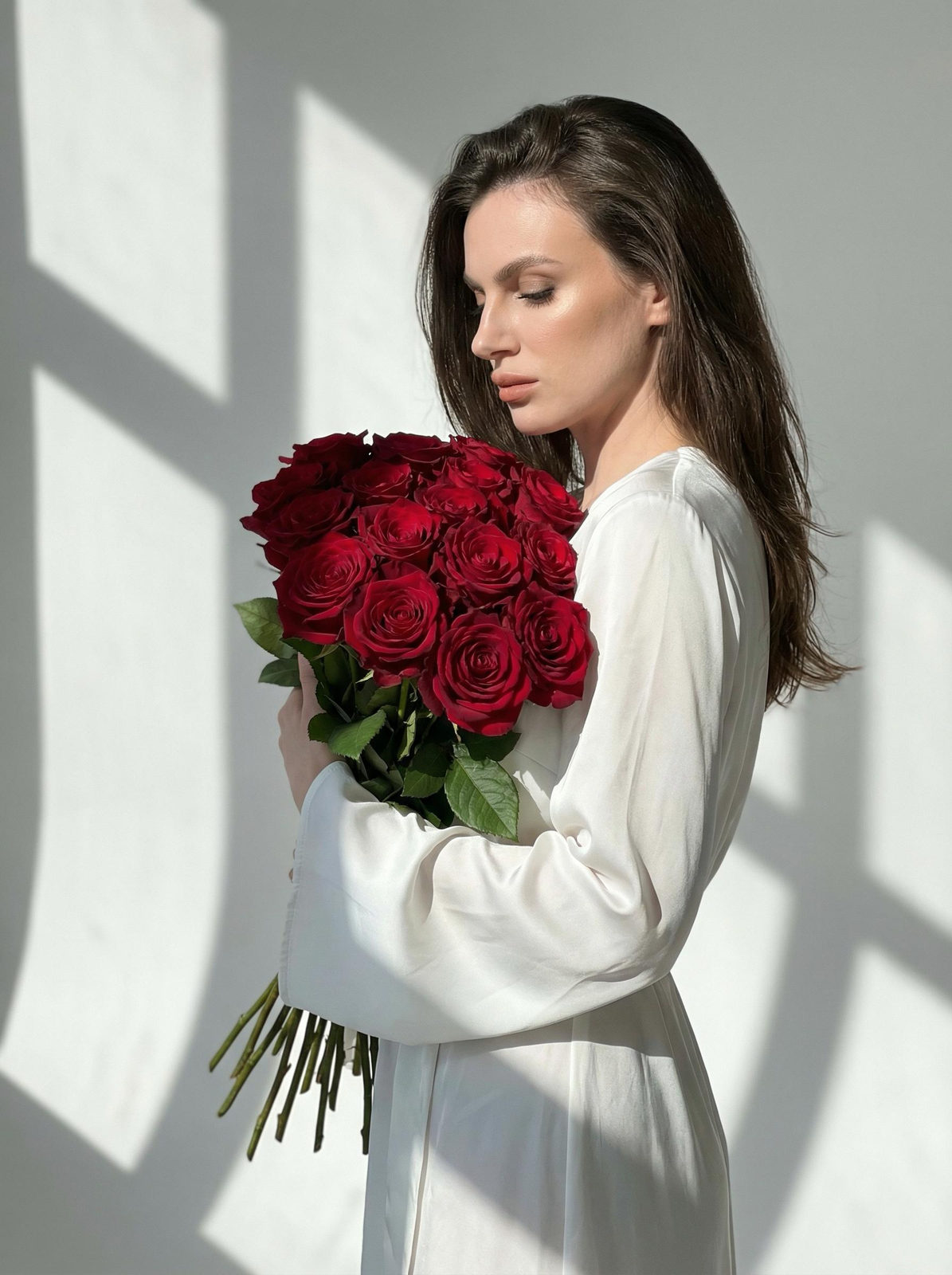 Woman holding a large bouquet of dark red roses in a bright studio, framed by geometric shadows.