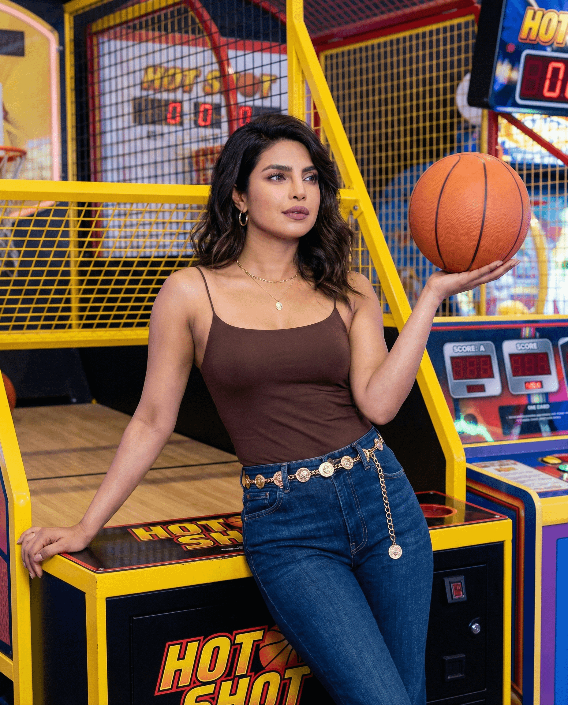 A confident woman in a chocolate tank top and denim jeans, leaning against an arcade machine with an orange basketball.