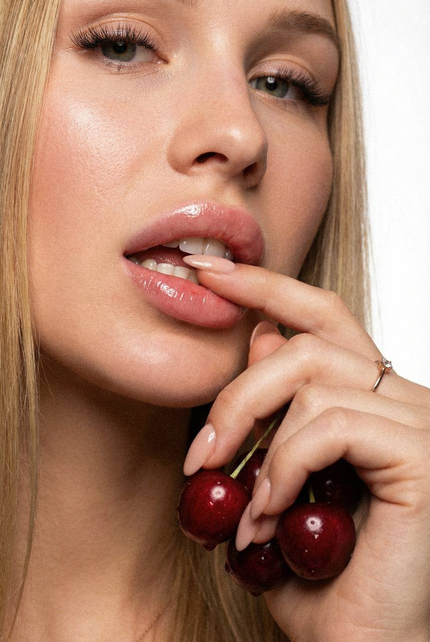 Extreme close-up of a woman's face with glossy lips, gently biting a fingertip while holding dark red cherries.