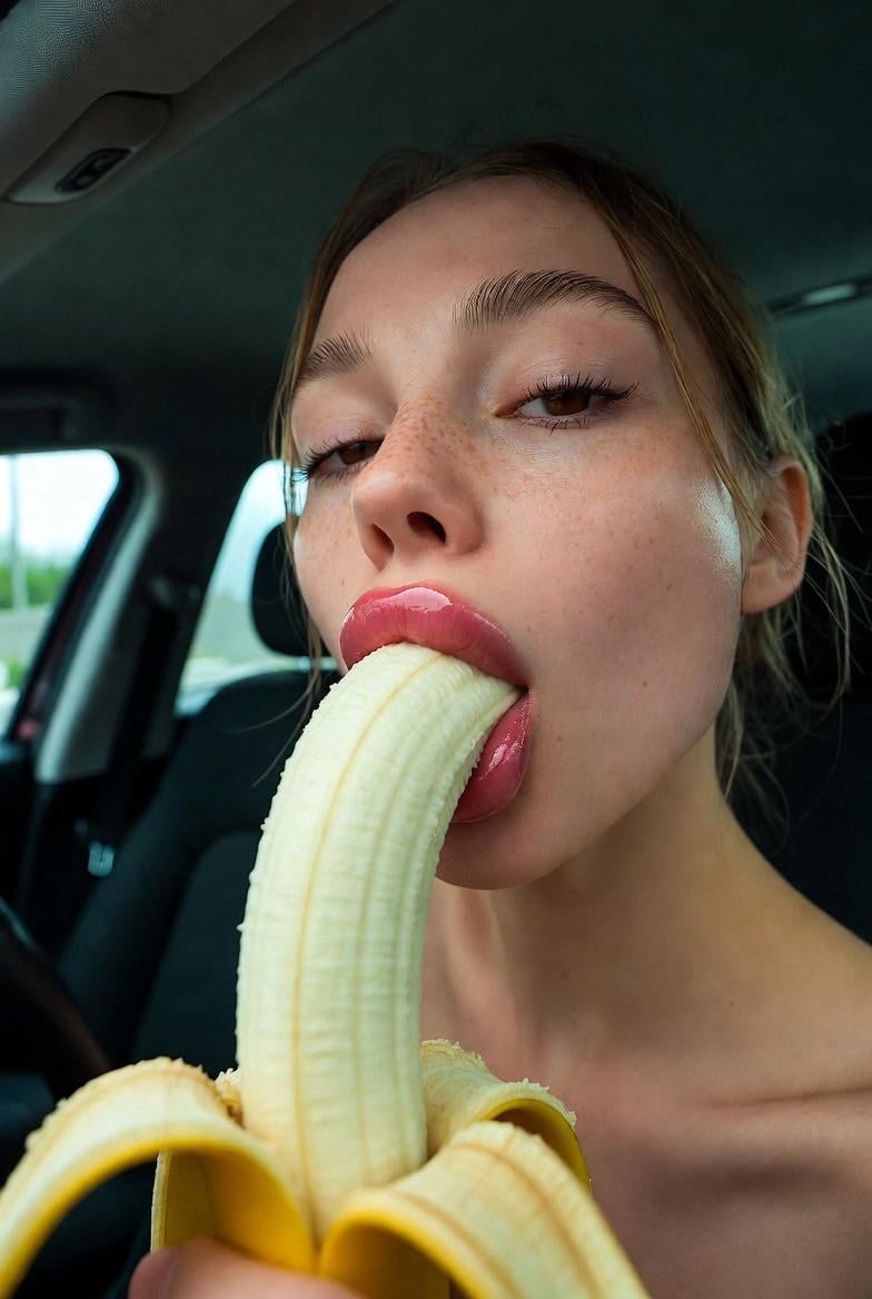 A close-up shot of a person in a car holding a banana, glossy pink parted lips, freckles visible, soft natural lighting.