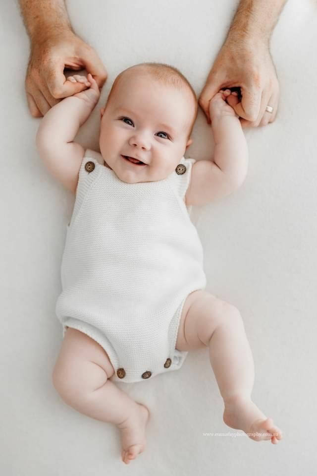 Overhead shot of a joyful baby smiling in a white knitted romper, held by adult hands on a soft white surface.