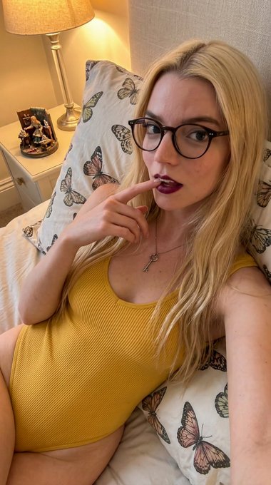 A woman with long wavy hair wearing a yellow bodysuit takes a playful selfie while lying in a cozy bedroom, softly lit by a bedside lamp.