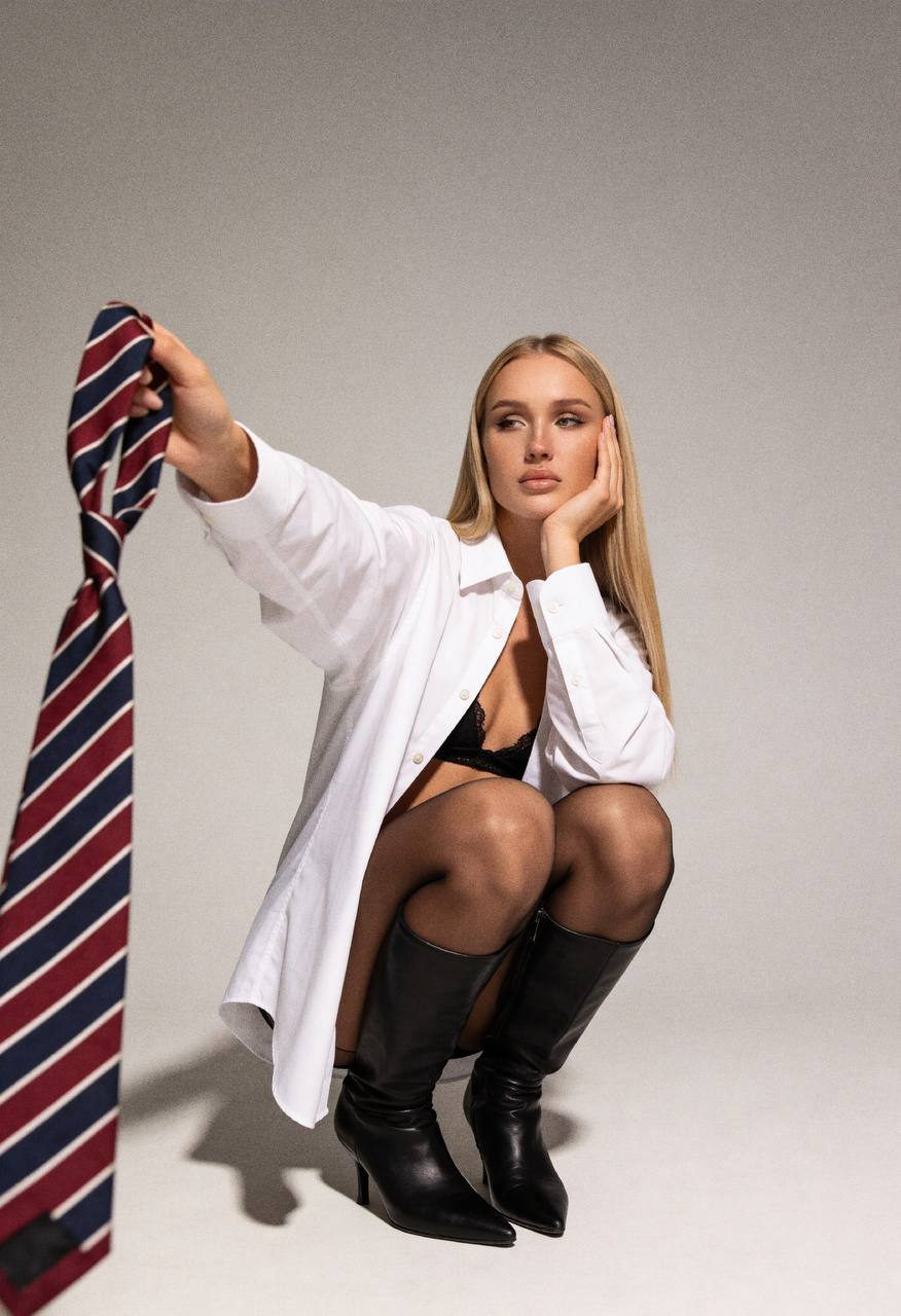 A young woman in a low squat, resting her head on her left hand, holding a striped tie with a neutral background.