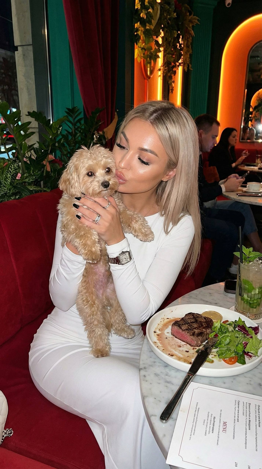 A woman kissing her maltipoo dog at a stylish cafe table, glowing skin and elegant decor around her.