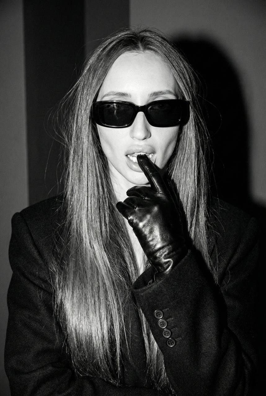 Close-up of a person in oversized sunglasses with shiny leather gloves, featuring a bold and relaxed pose.