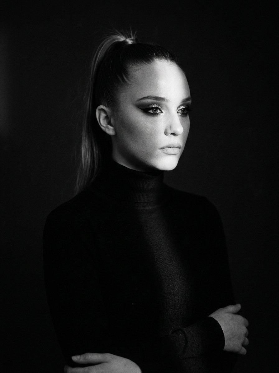 Dramatic waist-up portrait of a pensive woman in a black turtleneck, captured in dim studio light with bold makeup.